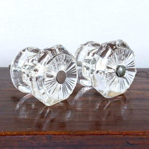 May include: Two clear glass cabinet knobs featuring an octagonal shape with a faceted, sunburst-like pattern radiating from the center. Each knob has a small, circular metal attachment point in the middle of the front face and a smooth, transparent finish.