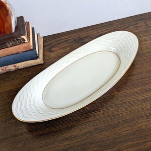 May include: An elongated oval ceramic serving dish with a cream-colored center and a textured basket-weave pattern along the rim. The edge of the dish is finished with a thin gold-colored trim, providing a subtle contrast to the white ceramic.