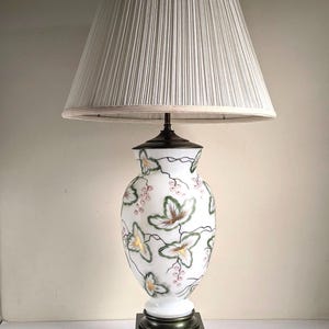 May include: A white ceramic table lamp with a floral design, featuring a pleated lampshade. The lamp base is a square, dark metal. The floral pattern includes green, yellow, and pink accents.