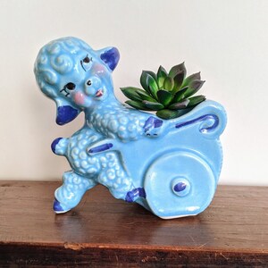 Op de afbeelding: Een lichtblauwe keramische lammetjesplanter met donkerblauwe accenten, met een groene vetplant. Het lam trekt een kar met een wiel. Het lam heeft roze wangen en een rode mond.