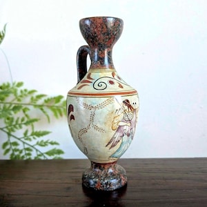 May include: A decorative ceramic vase with a handle, featuring a detailed illustration of a figure in classical attire. The vase has a speckled brown and beige base and top, with a cream-coloured body adorned with painted designs.