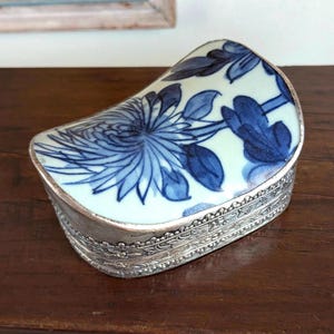 Puede incluir: Una caja de baratijas decorativa en forma de riñón. La tapa presenta un diseño floral azul y blanco, mientras que la caja tiene un marco de metal plateado con un patrón ornamentado. La caja se exhibe sobre una superficie de madera oscura.