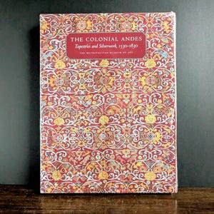 May include: A hardcover book titled "THE COLONIAL ANDES Tapestries and Silverwork, 1530-1830" from The Metropolitan Museum of Art. The book cover features a detailed, ornate pattern in red, yellow, and white.