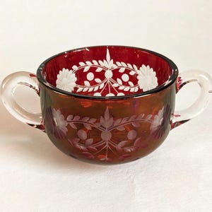 May include: A ruby red glass bowl with two clear handles. The bowl features etched floral and leaf patterns in white and clear glass. The rim is a deep red, and the bowl has a slight iridescent sheen.