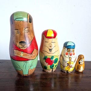 Può includere: Un set di quattro bambole a incastro in legno, ognuna dipinta con un design di animali stravaganti. La bambola più grande è un orso, seguita da un maiale, un musicista e un piccolo scoiattolo. Le bambole sono dipinte con colori vivaci.