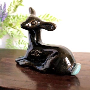 May include: A glossy, dark brown ceramic deer figurine in a resting pose. The figurine has a smooth, reflective surface with a touch of turquoise on the hooves. The text "ocono mts." is inscribed on the side.