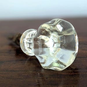 May include: A clear, vintage-style glass door knob with a rounded, irregular shape. The knob is mounted on a metal stem and sits on a dark wooden surface. The glass has a slight yellow tint.
