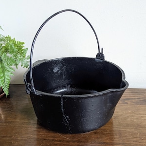 以下が含まれることがあります： 黒い鋳鉄製の鍋で、曲がった取っ手が付いています。鍋は口が広く開いており、底は丸みを帯びています。取っ手は鍋の側面に付いています。鍋は木製の表面に置かれています。
