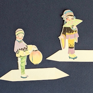 Könnte beinhalten: Zwei Papierauschnitte von Figuren in traditioneller asiatischer Kleidung. Eine Figur hält eine runde, gelbe Laterne. Die andere Figur hält einen Fächer. Beide Figuren stehen auf weißen Plattformen vor einem dunkelblauen Hintergrund. Die Figuren tragen bunte Kleidung.