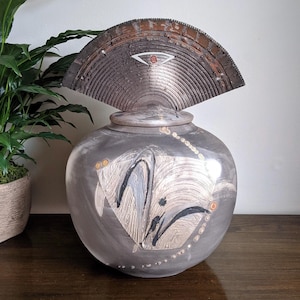 May include: A decorative ceramic vase with a fan-shaped lid. The vase is primarily grey with abstract black and white designs. The lid is a darker, textured brown. The vase sits on a wooden surface.