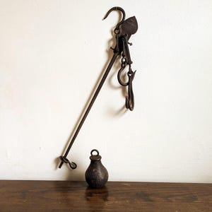 May include: An antique, rusty metal hanging scale with a long arm, multiple hooks, and a small, pear-shaped weight. The scale is suspended against a white wall and rests on a wooden surface. The scale is likely used for measuring weight.