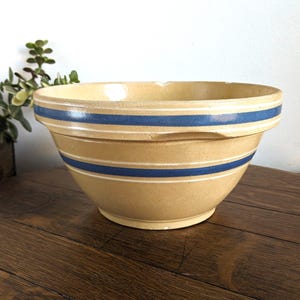 May include: A tan ceramic mixing bowl featuring two horizontal blue stripes encircling the upper and middle sections. The bowl has a wide, flared rim with a notched edge detail and a smooth, rounded base, showcasing a classic kitchen design.