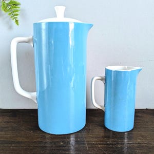 Könnte beinhalten: Eine weiße Keramik-Tasse mit schwarzem Henkel und dem Text "But First, Coffee" in fetter, schwarzer Schrift. Die Tasse steht vor einem verschwommenen Hintergrund, der eine gemütliche Umgebung suggeriert.
