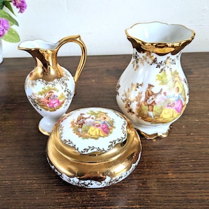 May include: A white porcelain set consisting of a small pitcher, a lidded jar, and a vase, all featuring gold-trimmed rims, ornate gold floral accents, and a central colorful illustration of two figures in a garden setting on each piece.
