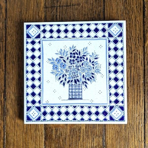 Puede incluir: Azulejo cerámico cuadrado con un diseño floral azul y blanco. La imagen central presenta un ramo de flores azules en un jarrón. El azulejo tiene un borde con patrón de diamantes y un marco cuadrado con un diseño similar.