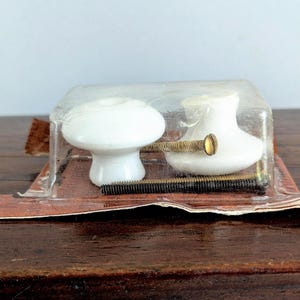 May include: Two white ceramic cabinet knobs in a clear plastic package. The knobs have a mushroom-like shape and are attached to brass screws. The package sits on a brown surface.