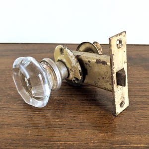 以下が含まれることがあります： 透明なファセットガラスのドアノブと、変色した真鍮製のラッチ機構を備えた、ヴィンテージのドアハードウェアセット。 金属部品は、エイジング加工が施され、経年劣化の兆候が見られます。 ハードウェアは木製の表面に置かれています。