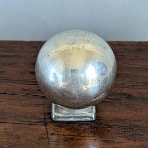 Pode incluir: Um puxador esférico de metal prateado com uma superfície polida e reflexiva. A esfera está montada sobre uma pequena base quadrada, criando uma peça única de ferragem com um topo arredondado e uma base plana e estável para fixação.