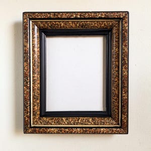 Pode incluir: Uma moldura retangular com interior preto e uma borda externa decorativa. A borda apresenta um padrão salpicado em tons de castanho, dourado e preto. A moldura foi projetada para conter uma fotografia ou obra de arte.