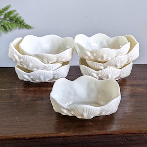 May include: A set of five off-white ceramic bowls, each featuring a moulded shell-like exterior with a scalloped rim. The bowls are arranged in two stacks of two with one bowl placed in the foreground, all showcasing a glossy finish and textured surface.