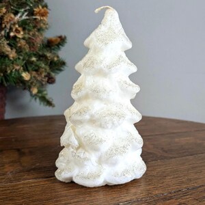 Può includere: Una candela bianca a forma di albero con un design strutturato a strati che ricorda i rami di pino. La superficie della candela è cosparsa di fine glitter dorato scintillante e un singolo stoppino sottile è visibile sulla punta.