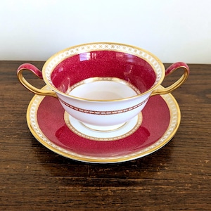 May include: A two-handled bouillon cup and saucer set featuring a deep magenta red band with a fine gold filigree pattern. The interior of the cup and the center of the saucer are white, accented by gold rims and delicate gold decorative borders.