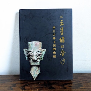 May include: A book with a black cover featuring gold Chinese characters and a bronze sculpture of a face. The sculpture has large ears and a detailed face. The book is on a wooden surface.