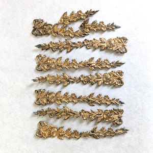 May include: A collection of seven elongated, gold-toned metal decorative appliqués arranged in rows. Each piece features a detailed relief design of floral garlands, leaves, and a bow at one end, with a tapered, pointed shape at the opposite end.