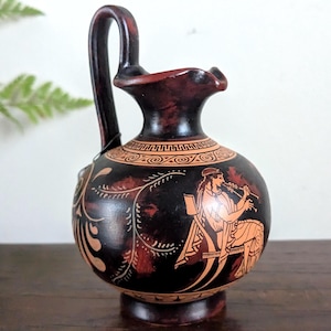 May include: A dark brown ceramic jug with a handle and a spout. The jug features a painted scene of a person playing a flute, with decorative patterns around the neck and body. The jug is likely a decorative item.