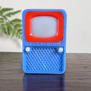 Puede incluir: Un pequeño televisor de juguete azul con un marco rojo alrededor de la pantalla. La palabra "TELEVISION" está impresa en letras blancas. La parte inferior del juguete tiene un área de altavoz con diseño de diamantes.