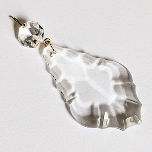 May include: A clear crystal chandelier prism with a faceted top and a teardrop-shaped bottom. The prism is connected to a gold-colored metal wire and a small, octagonal crystal. The background is white.