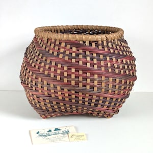 May include: A round woven basket featuring a pattern of tan, deep red, and dark brown splints. The basket has a thick, light tan rim and a wide, rounded body that tapers slightly toward the top. A small rectangular business card is placed nearby.