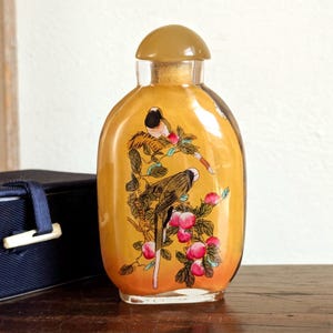 May include: A small, clear glass bottle with a yellow interior and a brown stopper. The bottle features a painted design of two birds perched on a branch with red fruit. The bottle is oval-shaped and sits on a wooden surface.