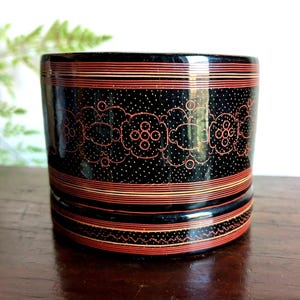 May include: A cylindrical black lacquer container featuring intricate red and gold line work. The exterior is decorated with horizontal bands, a central repeating floral pattern, and fine stippled dots, creating a detailed geometric design.