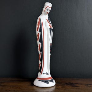 May include: A white ceramic figurine of a woman with a long dress. The figure has black and red decorative accents. The figurine is standing on a white base and is set against a dark background.