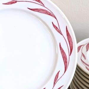 May include: White ceramic plates with a red leaf design around the rim. The plates are stacked, with one plate in focus, showcasing the detailed red leaf pattern. The plates are clean and simple, suitable for everyday use.