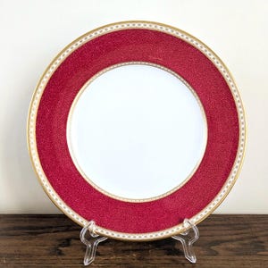 May include: A round porcelain dinner plate featuring a solid white center surrounded by a wide, textured crimson red border. The rim is decorated with a delicate gold and white geometric pattern, finished with a thin gold band along the edge.