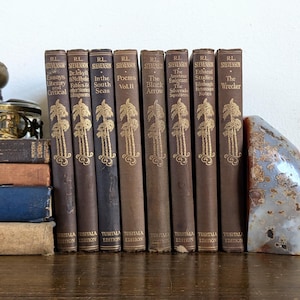 May include: A row of vintage books with brown spines and gold lettering. The titles include works by R.L. Stevenson, such as "The Black Arrow" and "Poems Vol. II." The books are displayed on a wooden surface.