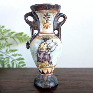 May include: A ceramic amphora vase featuring a dark mottled finish on the handles and base. The central body displays a painted figure playing a lyre, framed by a Greek key pattern above and a floral motif on the neck, with a small metal tag attached.