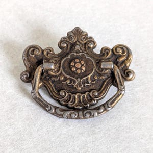 May include: A bronze-toned metal drawer pull featuring an ornate, symmetrical backplate with scrollwork details and a central circular floral motif. A curved, hinged drop handle with engraved patterns is attached to the backplate, creating a decorative hardware piece with a weathered, aged finish.