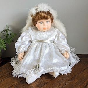 Puede incluir: Muñeca de porcelana vestida de ángel, con un vestido de satén blanco con ribete de encaje y un halo de plumas. La muñeca tiene ojos azules y cabello castaño. Artículo decorativo.