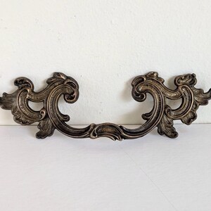May include: A bronze-toned metal drawer pull featuring an ornate, symmetrical design with scrolling leaf-like patterns. The hardware has a dark, antiqued finish with highlights on the raised edges, showcasing a curved, wide shape with two mounting points.