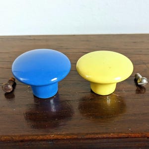 May include: Two cabinet knobs, one blue and one yellow, with matching screws. The knobs have a rounded, mushroom-like shape. The knobs are on a dark wooden surface.