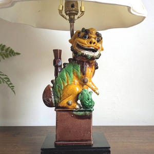 May include: A ceramic table lamp featuring a detailed, colorful Foo Dog figurine. The dog is yellow, green, and brown, with a smiling expression. The lamp has a cream-colored lampshade and sits on a black base.