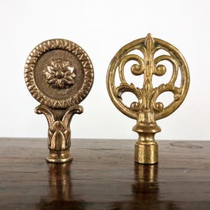 May include: Two brass lamp finials. The left finial is a circular disc with a central flower relief and a serrated border. The right finial features an openwork scroll design within a circular frame. Both have a cylindrical base for attachment.
