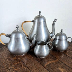 Puede incluir: Un juego de té de peltre vintage, que incluye una tetera, una crema, un azucarero y otra tetera. La tetera de la izquierda tiene un asa tejida. El juego se exhibe sobre una superficie de madera.