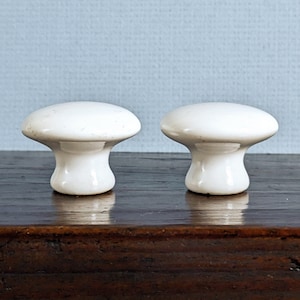 May include: Two white ceramic mushroom-shaped cabinet knobs. The knobs have a rounded top and a flared base. They are set against a light blue background and sit on a dark wooden surface. These knobs are ideal for adding a touch of vintage charm to furniture.
