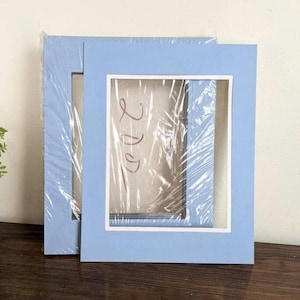 May include: Two light blue picture frames with white matting are stacked on a wooden surface. The frames are wrapped in clear plastic. The frames have a rectangular opening for the photo.
