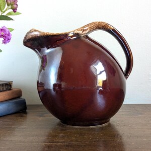 May include: A glossy, dark brown ceramic pitcher with a rounded, bulbous body and a curved handle. The rim features a textured, lighter brown drip glaze detail. The surface is smooth and reflective, showing light highlights across the curved form.