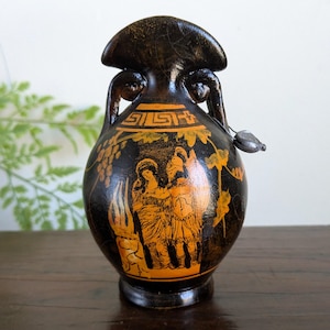 May include: A black ceramic amphora vase featuring orange-toned illustrations of two figures in classical attire. The neck displays a geometric Greek key pattern, while the body is decorated with grapevines and a small metallic charm attached to the handle.